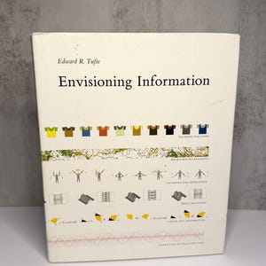 May include: A white hardcover book titled Envisioning Information by Edward R. Tufte. The cover features various graphic illustrations including small t-shirt icons, semaphore signals, geometric shapes, and diagrams labeled with text like Escaping Flatland.
