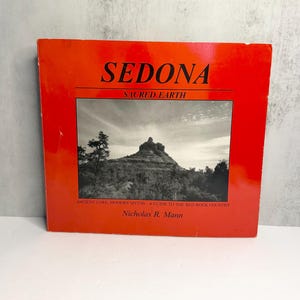 May include: A square book titled Sedona Sacred Earth by Nicholas R. Mann. The cover is bright red with black text and features a centered black and white photograph of a rocky mountain landscape under a cloudy sky.