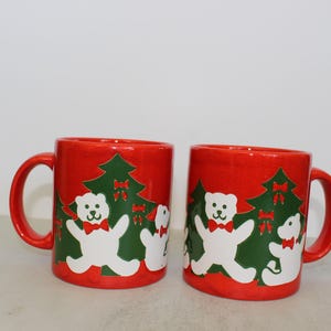 Puede incluir: Dos tazas de cerámica roja con temática navideña. Cada taza presenta un oso polar blanco con una pajarita roja de pie frente a un árbol de Navidad verde con adornos rojos.