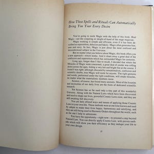 May include: Open book with text titled "How These Spells and Rituals Can Automatically Bring You Your Every Desire." The page has paragraphs of text with the number 27 at the bottom. The book is open on a white surface.