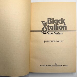 May include: Open book with the title "The Black Stallion and Satan" by Walter Farley. The title is in a stylized black font with a decorative swirl. The book is published by Random House, New York.