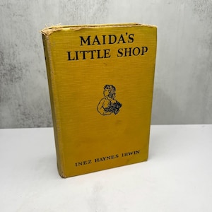 May include: A vintage book titled "Maida's Little Shop" by Inez Haynes Irwin. The book has a mustard yellow cover with black text and a small illustration of a girl. The book's spine shows wear and tear.