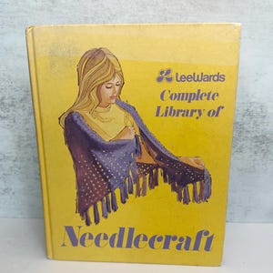 May include: A yellow hardback book titled Leewards Complete Library of Needlecraft. The cover features a printed illustration of a person with long blonde hair wearing a purple fringed shawl over a yellow top, with purple text throughout.