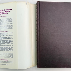 May include: An open book titled "The Mystic Grimoire of Mighty Spells and Rituals" by Frater Malak. The book has a dark brown cover and white pages with text. The left page has text in purple and blue.