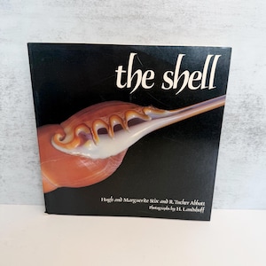 May include: A hardback book titled The Shell with a black cover featuring a large, detailed photograph of an orange and white seashell. The text Hugh and Marguerite Stix and R. Tucker Abbott photographs by H. Landshoff is printed in white.