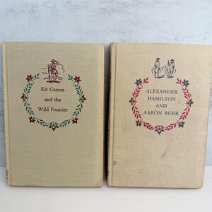May include: Two vintage hardcover books with beige covers. The book on the left is titled "Kit Carson and the Wild Frontier" and the book on the right is titled "Alexander Hamilton and Aaron Burr". Both books feature illustrations and text in red and green.
