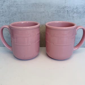 May include: Two matching dusty pink ceramic mugs featuring a raised, embossed basket-weave pattern around the center of the body. Each mug has a smooth, rounded handle on the side and a wide, circular rim, presenting a uniform, solid-colored finish.