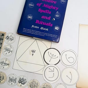 May include: A blue book titled "Mystic Grimoire of Mighty Spells and Rituals" by Frater Malak. The image also shows several circular and geometric illustrations with symbols and numbers, and a white paper with a triangle and a flame.