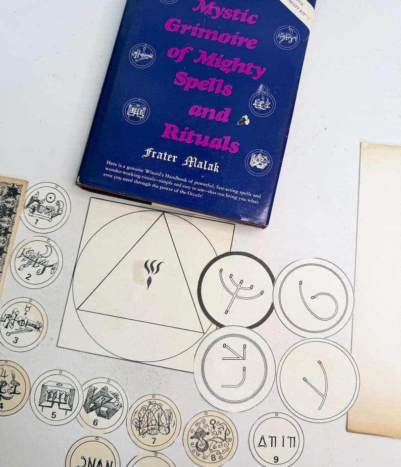 May include: A blue book titled "Mystic Grimoire of Mighty Spells and Rituals" by Frater Malak. The image also shows several circular and geometric illustrations with symbols and numbers, and a white paper with a triangle and a flame.