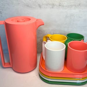 May include: A coral-colored pitcher with a handle and lid, next to a tray with four mugs in white, yellow, green, and coral. The mugs sit on a stack of colorful trays in orange, yellow, white, and green. A retro-style drinkware set.