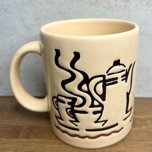 May include: Cream ceramic coffee mug featuring a black line drawing of a coffee pot, cup, and steam. The mug has a curved handle and a brown base. The design is simple and graphic, suitable for hot beverages.