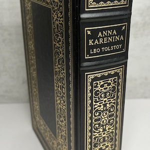 May include: A hardback book titled "ANNA KARENINA" by Leo Tolstoy. The book has a black cover with gold decorative filigree and lettering. The spine also features gold lettering and decorative elements.