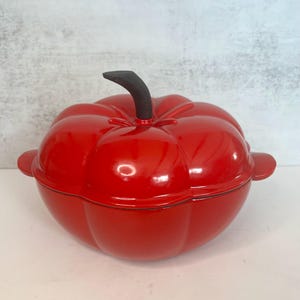 May include: A bright red, pumpkin-shaped cast iron Dutch oven with a black stem handle. The pot has a glossy finish and two side handles. This cookware is ideal for cooking and serving soups, stews, and other dishes.