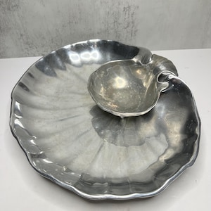 May include: A silver-colored serving dish with a seashell design. The dish has a large, scalloped-edge plate with a smaller, shell-shaped bowl attached. The surface has a textured, ridged pattern.