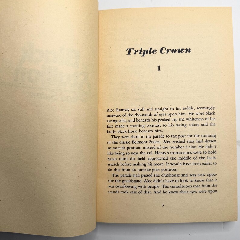 May include: Open book pages with the title "Triple Crown" and the number "1" at the top. The text describes a horse race, mentioning Alec Ramsay, racing silks, and the Belmont Stakes. The page has a cream color.