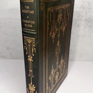 May include: A hardcover book titled "The Alcestiad" by Thornton Wilder. The book has a dark green cover with gold decorative accents and lettering. The spine reads "First Edition" and "The Franklin Library".
