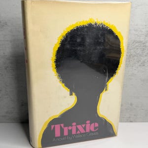 May include: Vintage paperback book titled "Trixie" by Wallace Graves. The cover features a silhouette of a person with an afro, outlined in yellow, with the title in pink. The book has a cream-coloured background.