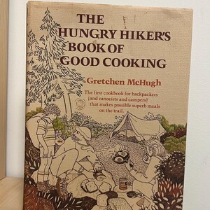 May include: A vintage cookbook, "The Hungry Hiker's Book of Good Cooking" by Gretchen McHugh. The cover illustration depicts a camping scene with people cooking and a tent. It's a guide for backpackers and campers, with text on the cover.