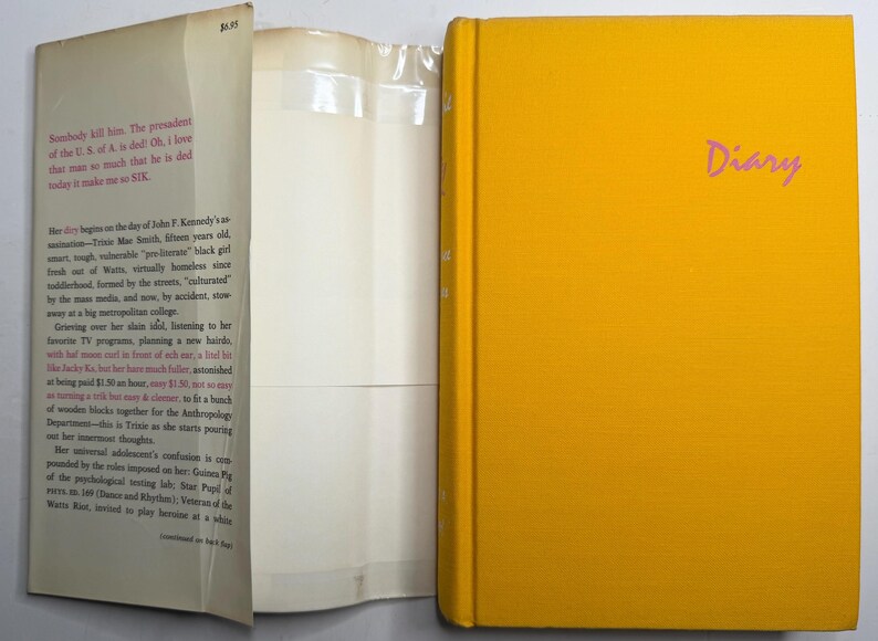 May include: A yellow hardcover book titled "Diary" in pink script. The book is open, showing white pages and text on the inside cover. The spine of the book is also yellow. The book's cover is plain.