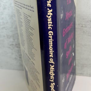 May include: A vintage book titled "The Mystic Grimoire of Mighty Spells and Rituals" by Peter Malak. The book has a blue spine with white text and a cream-colored cover. The book's title is also printed on the cover in pink.