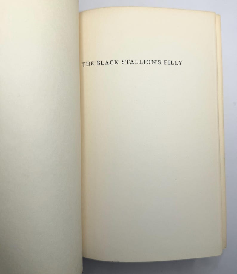 May include: Open book with the title "THE BLACK STALLION'S FILLY" printed in black on a cream-colored page. The book is open to a double-page spread, with the text centered on the right-hand page.