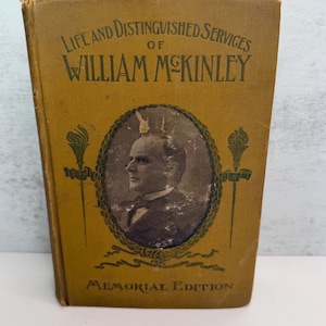 May include: A hardcover book titled Life and Distinguished Services of William McKinley Memorial Edition. The mustard-yellow cover features a central oval portrait of William McKinley, flanked by green torch illustrations and dark green text.
