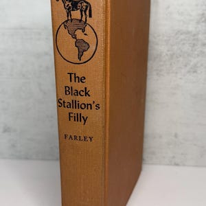May include: A vintage hardcover book titled "The Black Stallion's Filly" by Farley. The book has a brown cover with a black illustration of a horse above a globe. The publisher's name, Random House, is at the bottom.