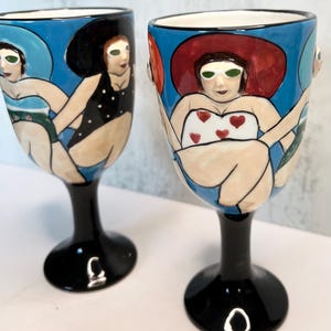 May include: Two hand-painted ceramic wine glasses. Each glass features a black stem and base, with a blue rim and a painted illustration of a woman in a swimsuit and sunglasses. One woman wears a black polka dot swimsuit, the other a white swimsuit with red hearts.