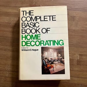 以下が含まれることがあります： 「THE COMPLETE BASIC BOOK OF HOME DECORATING」というタイトルのヴィンテージ本。クリーム色の表紙で、タイトルは黒と緑の文字で書かれています。表紙には、装飾されたリビングルームの写真が掲載されています。William E. Hague編集。