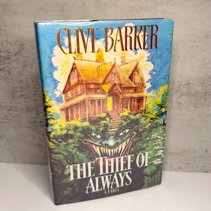 May include: A hardcover book titled The Thief of Always by Clive Barker. The cover features a golden illustration of a large house set against a blue sky, with a green, sharp-toothed creature face emerging from the foliage beneath the house.