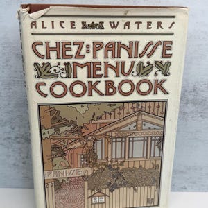 May include: A hardcover book titled Chez Panisse Menu Cookbook by Alice Waters. The off-white cover features brown text and a detailed line-art illustration of a building exterior with a fence, trees, and a sign reading Panisse.