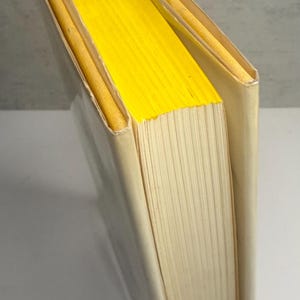 May include: A hardback book with a cream-coloured cover and bright yellow page edges. The book is slightly angled, revealing the yellow pages and the spine. The pages are off-white.