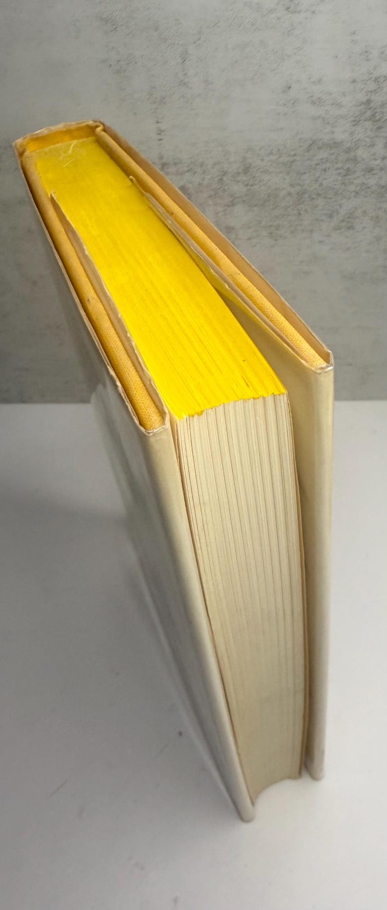 May include: A hardback book with a cream-coloured cover and bright yellow page edges. The book is slightly angled, revealing the yellow pages and the spine. The pages are off-white.