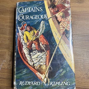 May include: Vintage hardcover book titled "Captains Courageous" by Rudyard Kipling. The cover features an illustration of a fishing boat with men and fish, set against a blue ocean backdrop. The book's title is in a stylized font.