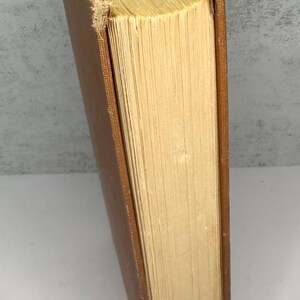 May include: A vintage book with a brown cover and aged, yellowed pages. The book's spine is visible, showing the binding and the wear and tear of time. The pages are thick and have a rough texture, suggesting age and use.
