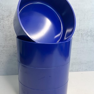 May include: A stack of three cobalt blue cylindrical containers, with a fourth container partially placed on top. The containers have a smooth, glossy finish and are set against a light gray background.