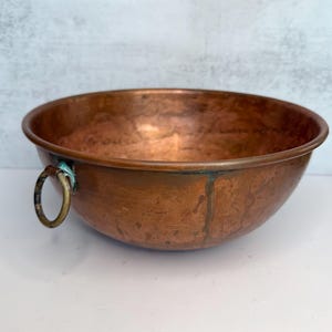 May include: A copper bowl with a rounded shape and a tarnished patina. The bowl has a brass ring handle attached to the side. The interior of the bowl shows signs of use and age. The bowl is sitting on a white surface.