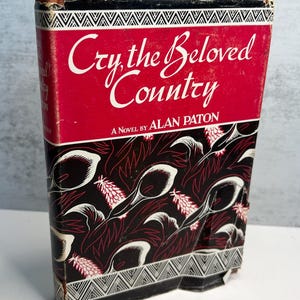 May include: A vintage book titled "Cry, the Beloved Country" by Alan Paton. The cover features a red background with white script and a black and white floral pattern. The book has a worn appearance.