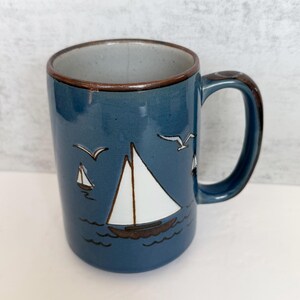 May include: A blue ceramic mug featuring a hand-painted white sailboat on the water with brown outlines. The design includes two small seagulls flying above the boat and a brown rim. The interior of the mug is a light speckled grey color.