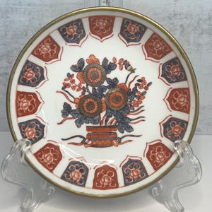 May include: A white ceramic plate with a gold rim and floral design. The plate features a central bouquet of orange and grey flowers, surrounded by red and blue floral patterns. The plate is displayed on a clear stand.