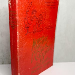 May include: A vintage cookbook titled "Mother's Day Everyday Cookbook" in a red cover with gold lettering. The cover features an illustration of a person cooking. The book is a facsimile of recipes and home remedies from 1950.