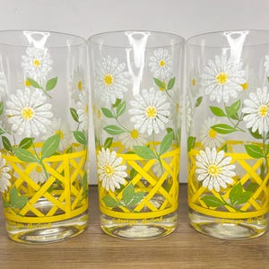May include: Three clear drinking glasses with a daisy floral design. The glasses feature white daisies with yellow centers, green leaves, and a yellow lattice pattern. The base of each glass has text.
