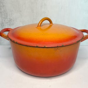 May include: A cast iron Dutch oven with a lid. The pot is painted in a gradient of orange and yellow, with a matching lid and handle. The pot has two side handles and a loop handle on the lid.