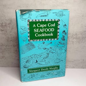 May include: A hardback book titled A Cape Cod SEAFOOD Cookbook by Margaret Deeds Murphy. The light blue cover features black line illustrations of various sea creatures, fishing nets, and buoys, with a dark green central text box.