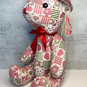 Peut inclure: Un ours en peluche patchwork avec un nœud rouge. L'ours est fait de carrés de tissu roses, blancs et verts avec des motifs de cœurs et de fleurs. Il a un œil en bouton noir et un nez rouge. Une étiquette est attachée à l'oreille.