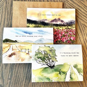 May include: A set of watercolour greeting cards with landscape scenes and inspirational quotes. The cards feature images of fields, trees, and mountains. One card reads, "I don't know what to say." Another says, "God is still writing your story."