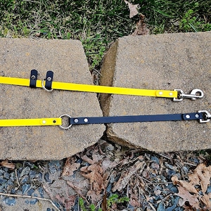 May include: Two dog leashes, one yellow and one black, are laid out on a stone surface. Both leashes have metal clips and a ring for attaching to a collar.