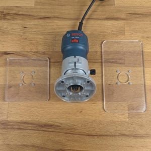 May include: A blue and silver Bosch GKF 125CE palm router with a black power cord, positioned between two clear acrylic base plates. The router features a circular base, a red power switch, and a metal housing with visible mounting screws.