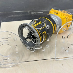 May include: A clear plastic router template with multiple holes and slots for a DEWALT 600 Series router. The router is yellow and black and is sitting on the template.