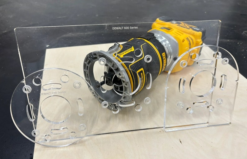 May include: A clear plastic router template with multiple holes and slots for a DEWALT 600 Series router. The router is yellow and black and is sitting on the template.
